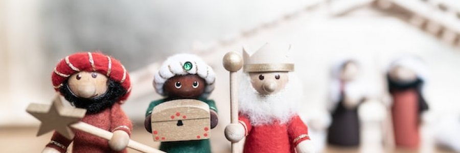 Image shows three wooded figures representing three wise men.  The leftmost figure is carrying a star on a stick, the middle a chest, and the rightmost an orb on a stick.  In the background, out of focus, is the nativity stable.